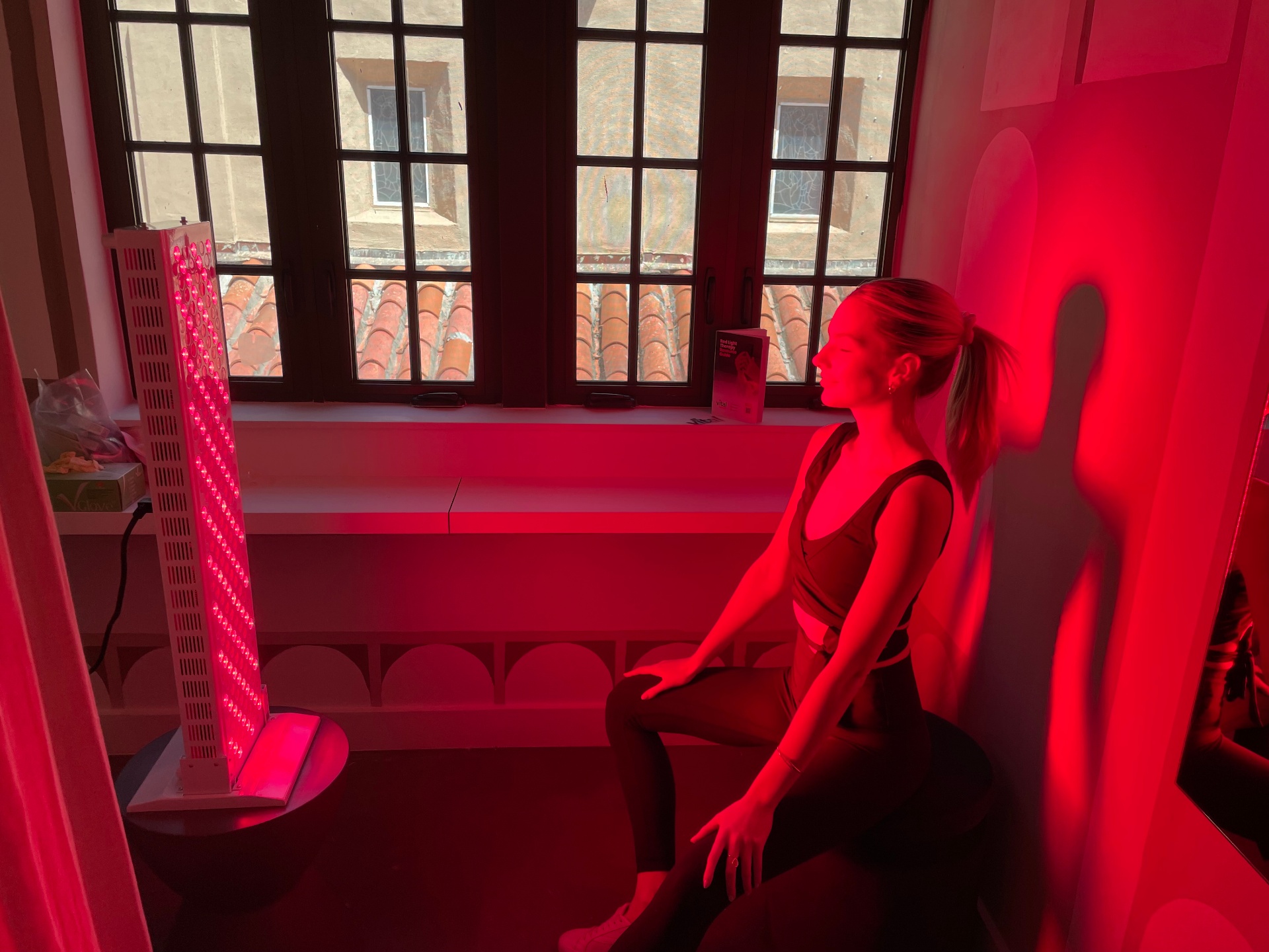 Woman seated near a tower-style red light therapy panel by a window